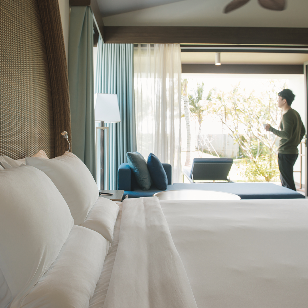 Luxurious hotel room with a comfortable bed, facing a private patio overlooking lush greenery. A person is visible in the background enjoying the outdoor space.