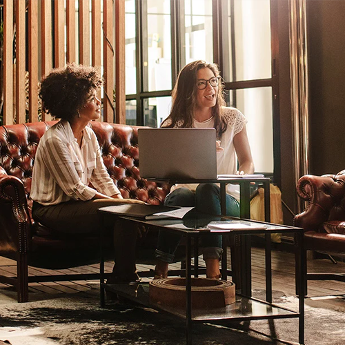 Two people sharing a laptop on leather couch in sunlit room with wooden accents