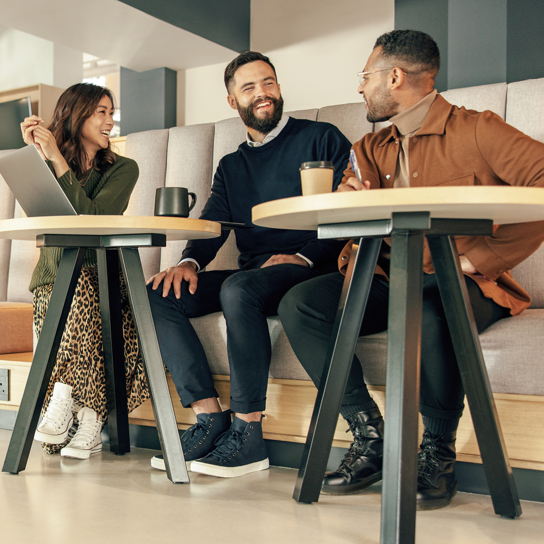 Three professionals having a meeting in a modern office space, seated on a couch with laptops and coffee.