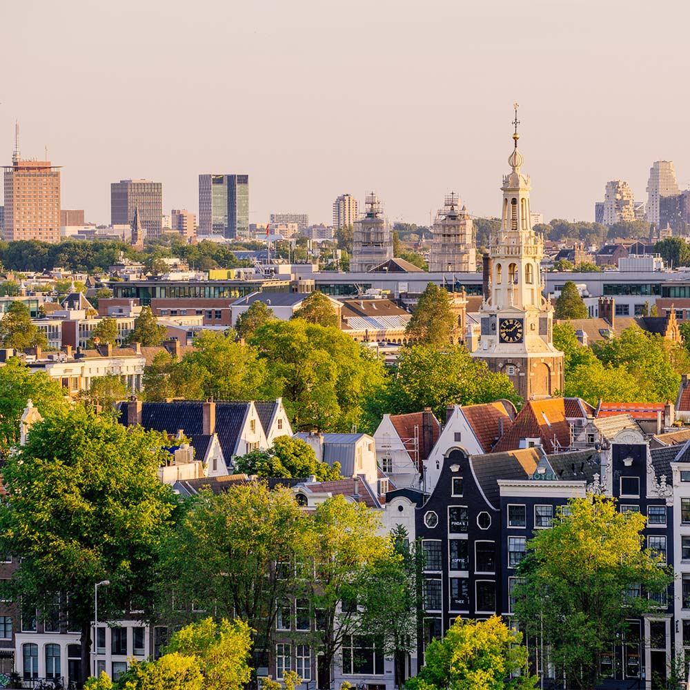 An urban panorama during the golden hour, which means the photo was taken shortly before sunset. The perspective is from an elevated point, with the viewer looking out over a densely built city with a lot of historical architecture. In the foreground, you see a series of traditional Dutch houses with characteristic stepped gables, which are close together. The facades of the houses are predominantly white, beige, and gray, with the occasional brick facade. The roofs are mostly red and orange tiles. The houses have large windows, some with white frames, and there are several dormer windows visible.