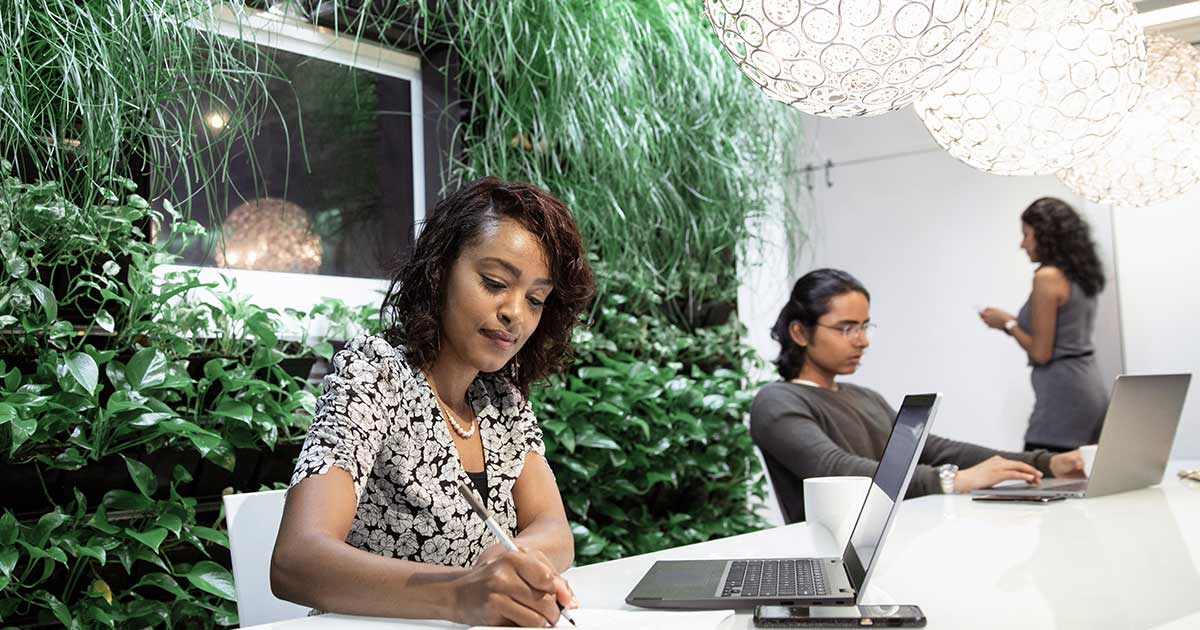 An office environment with a strong emphasis on greenery. In the foreground a woman is sitting at a white table working on a laptop. She appears to be writing something on a notepad with a pen.