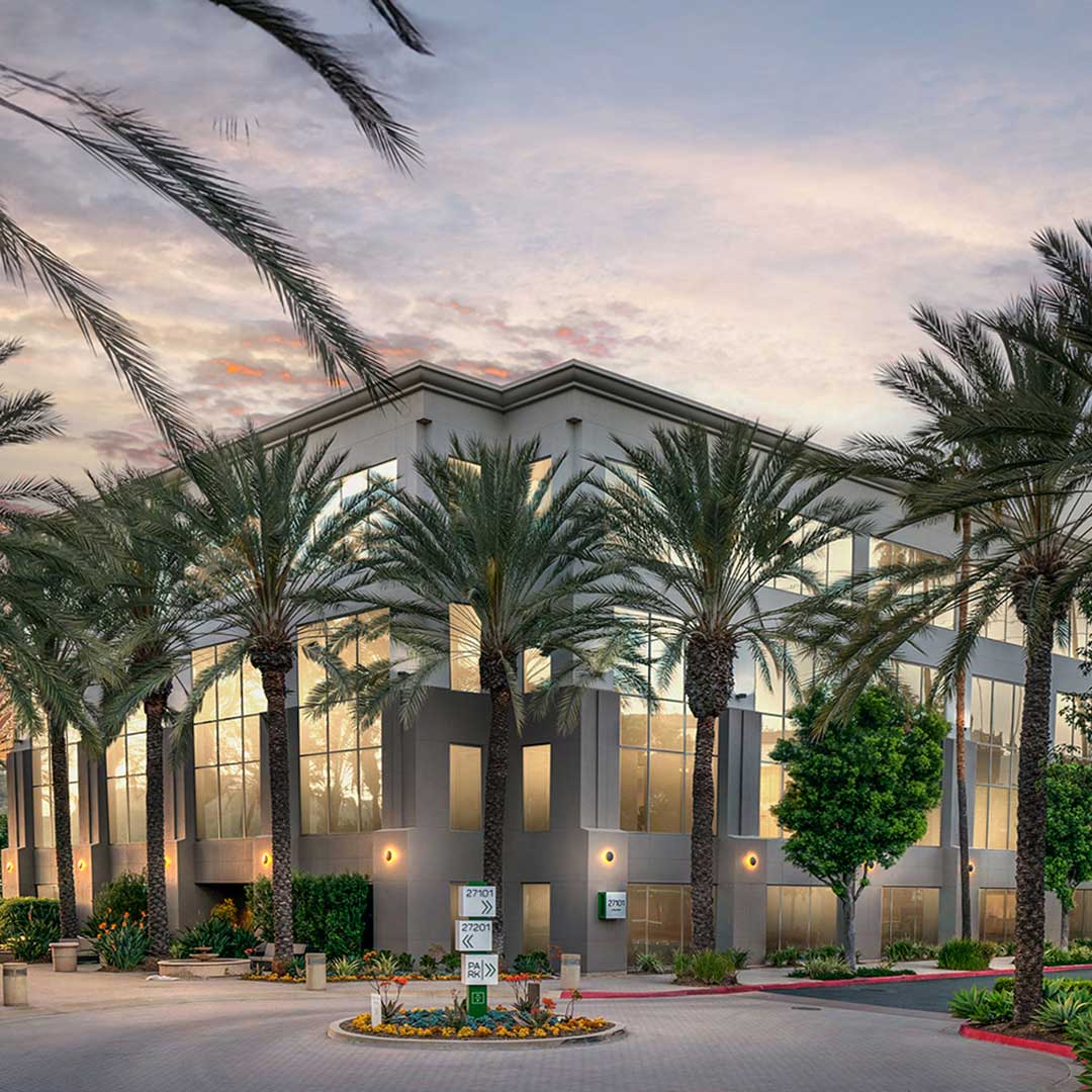 A large office building with glass windows, flanked by palm trees in the foreground, under a clear blue sky.