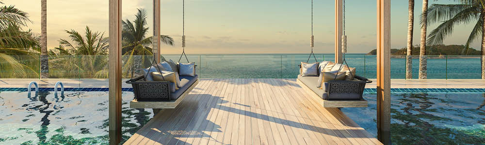 Panoramic view of a resort-style infinity pool overlooking the ocean. Two comfortable hanging daybeds are situated on a spacious wooden deck.