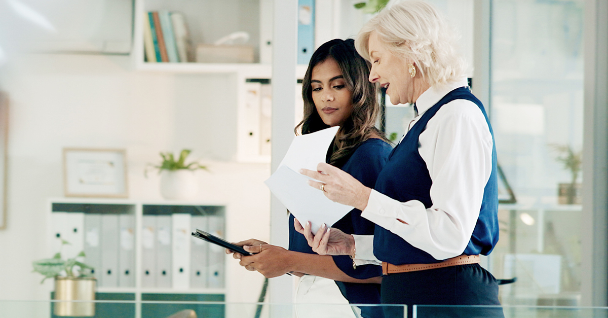 Two businesswomen collaborating, reviewing documents and using a tablet. This suggests a successful business partnership.