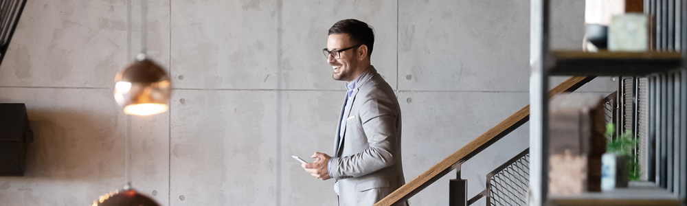 A smiling businessman in a suit walks down a modern staircase in a contemporary office building while looking at his phone.