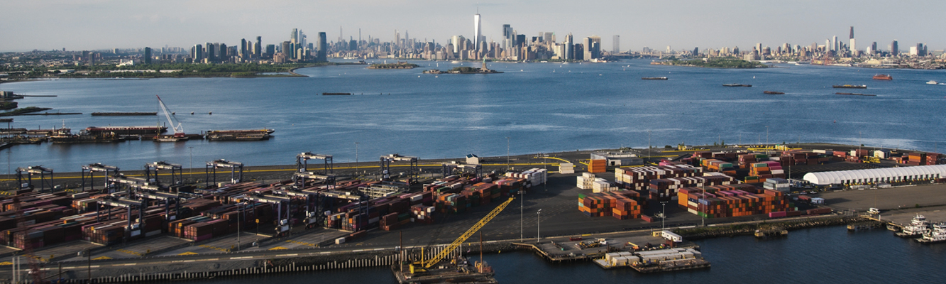 Aerial view of a New York City port showing many shipping containers, cranes, and the city skyline. 