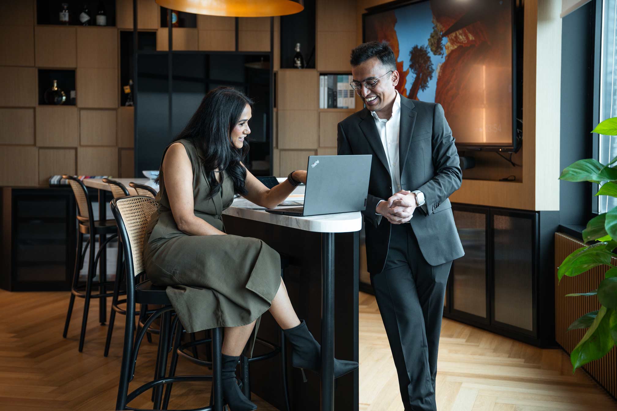 An indoor setting resembling a modern office or business lounge. It features two individuals, one seated (a woman in an olive green dress) working on a laptop and another standing (a man in a dark suit) holding a closed tablet or folder. The background includes a well-lit room with decorative items and a contemporary design aesthetic suitable for business activities.