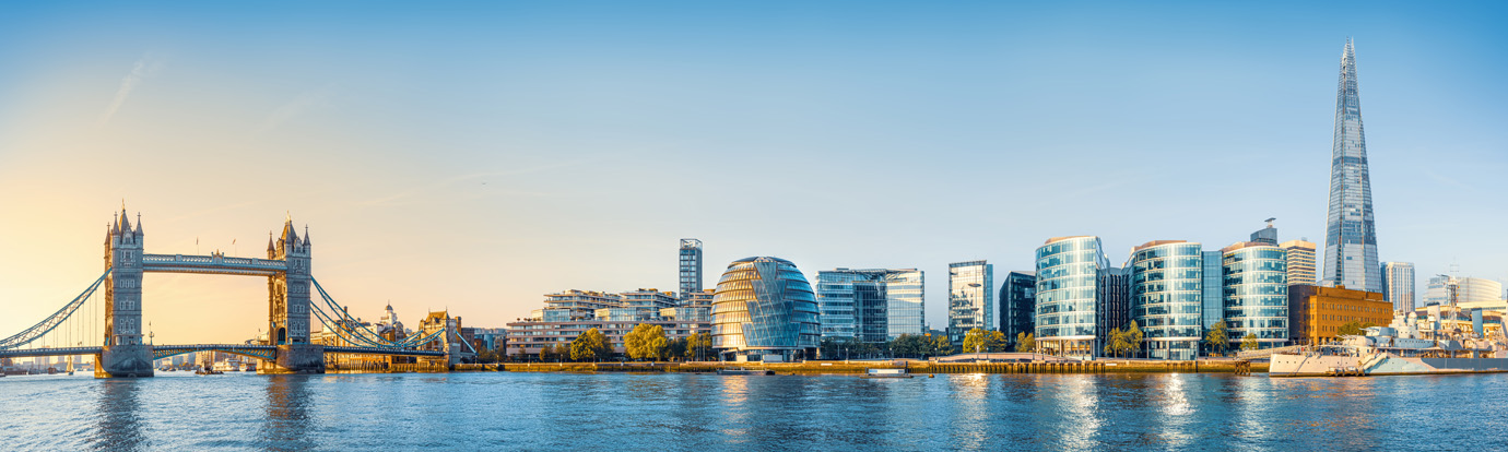 Panoramic view of the London skyline featuring Tower Bridge on the left and The Shard on the right, with modern buildings and the River Thames in the foreground under a clear blue sky.