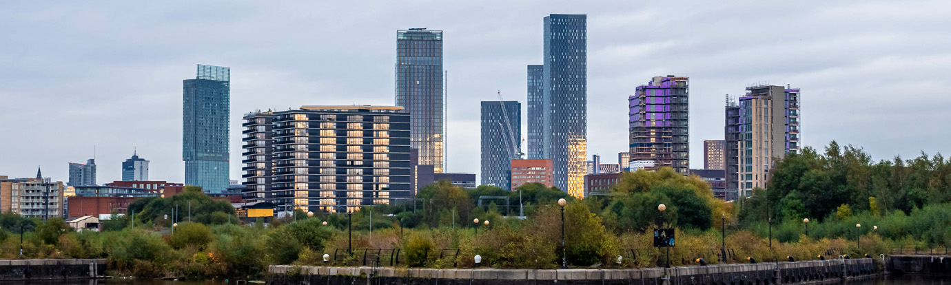A cityscape featuring a mix of modern high-rise buildings and mid-rise structures under a cloudy sky. Green trees and foliage are visible in the foreground, partially obscuring the lower parts of the buildings.