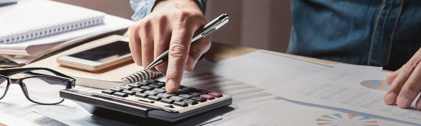 Close-up of a person using a calculator on a cluttered desk with documents, a smartphone, glasses, and a notepad. The person is holding a pen and appears to be in the middle of calculations or financial work.
