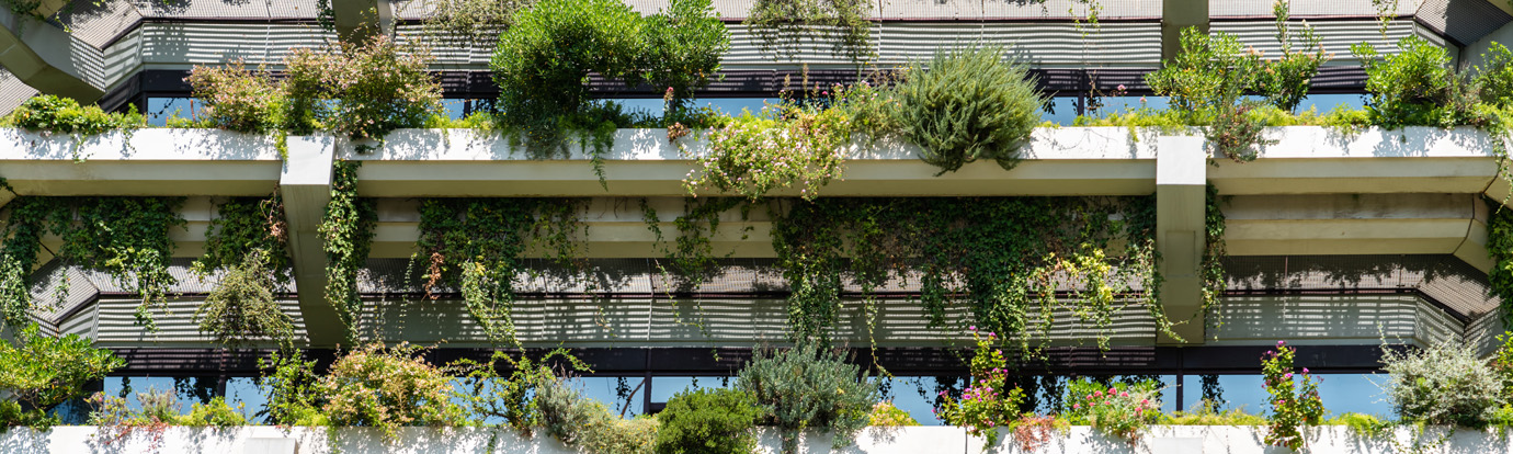 A modern building facade adorned with lush green plants and foliage cascading over white horizontal beams and balconies. The greenery contrasts with the building's clean architectural lines, creating a blend of nature and contemporary design.