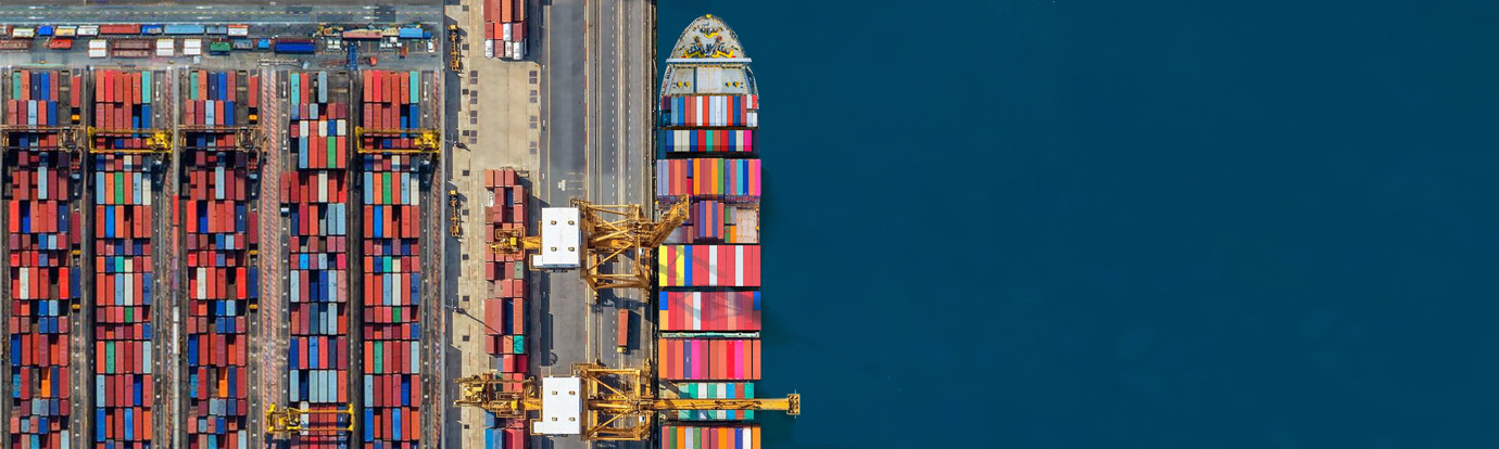 Aerial view of a cargo ship docked at a port, surrounded by numerous colorful shipping containers both on the ship and stacked on the ground. Yellow cranes are positioned along the dock, loading and unloading the containers.