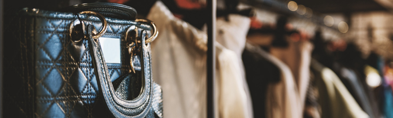A close-up of a stylish quilted handbag with metal hardware hanging on a rack in a clothing store. In the background, there are various garments hanging, slightly out of focus, giving the setting a chic and fashionable ambiance.