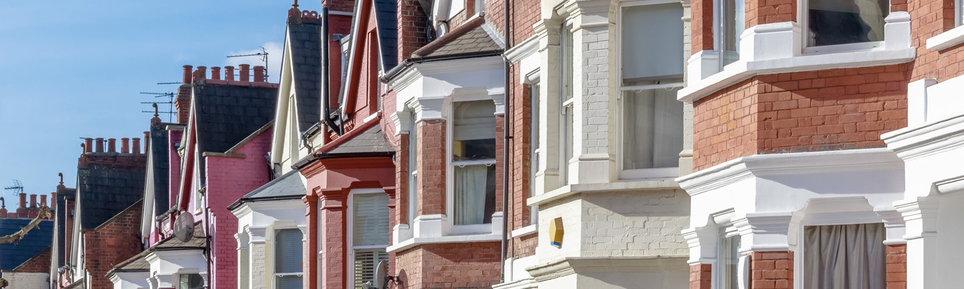A row of traditional brick townhouses with white trim and variously colored facades on a sunny day. The homes have bay windows and chimneys rising from the rooftops.