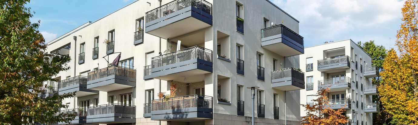 A modern apartment complex with multiple balconies, featuring light-colored exterior walls and blue railings. The buildings are surrounded by trees with green and autumnal foliage under a partly cloudy sky.