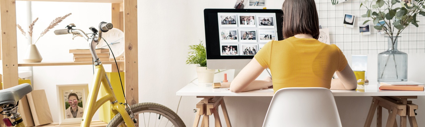 A person wearing a yellow shirt is sitting at a white desk and working on a computer with a collage of photos on the screen. There is a yellow bicycle beside the desk, and the room features plants, shelves with books, and various decorative items.
