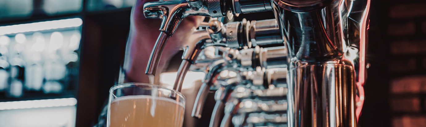 A row of beer taps dispensing a light-colored beer into a glass at a bar. The scene is illuminated with soft, ambient lighting, highlighting the metal taps and creating a warm, inviting atmosphere.