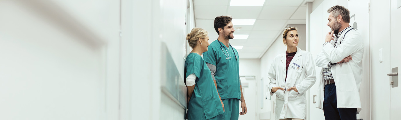 Four medical professionals, two men and two women, in scrubs and lab coats, are standing in a hospital hallway having a discussion. The setting is well-lit with white walls and ceiling panels.