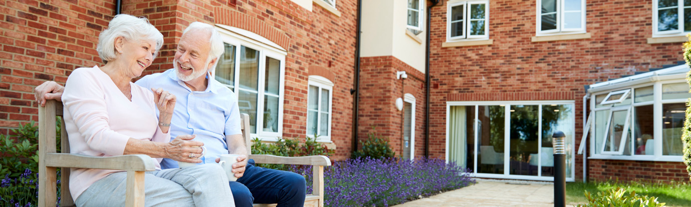 An elderly couple sits on a wooden bench outside a brick building, smiling and chatting. The woman is holding a cup, and the man has his arm around her. They are surrounded by a neatly manicured garden with purple flowers and a pathway leading to the building.