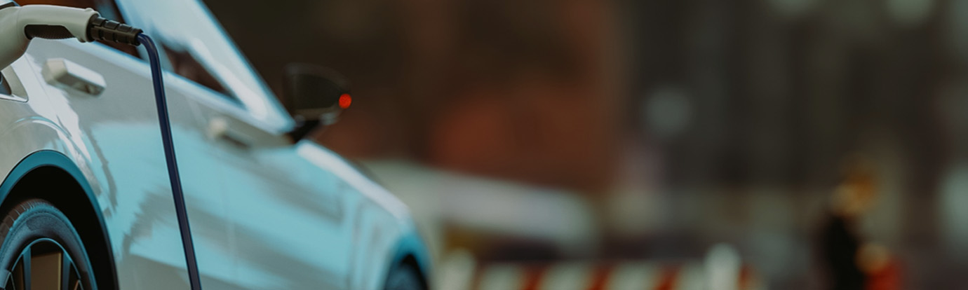A close-up of an electric car being charged. The blue charging cable is plugged into the car's port on the driver's side. The car is white, and the background is blurred, making the vehicle and the charging cable the clear focus of the image.