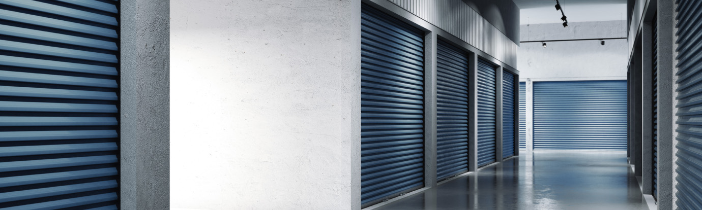A well-lit storage facility corridor with rows of closed blue metal rolling doors on either side. The floor is clean and shiny, and the walls are white. Ceiling lights are visible, providing consistent illumination throughout the hallway.