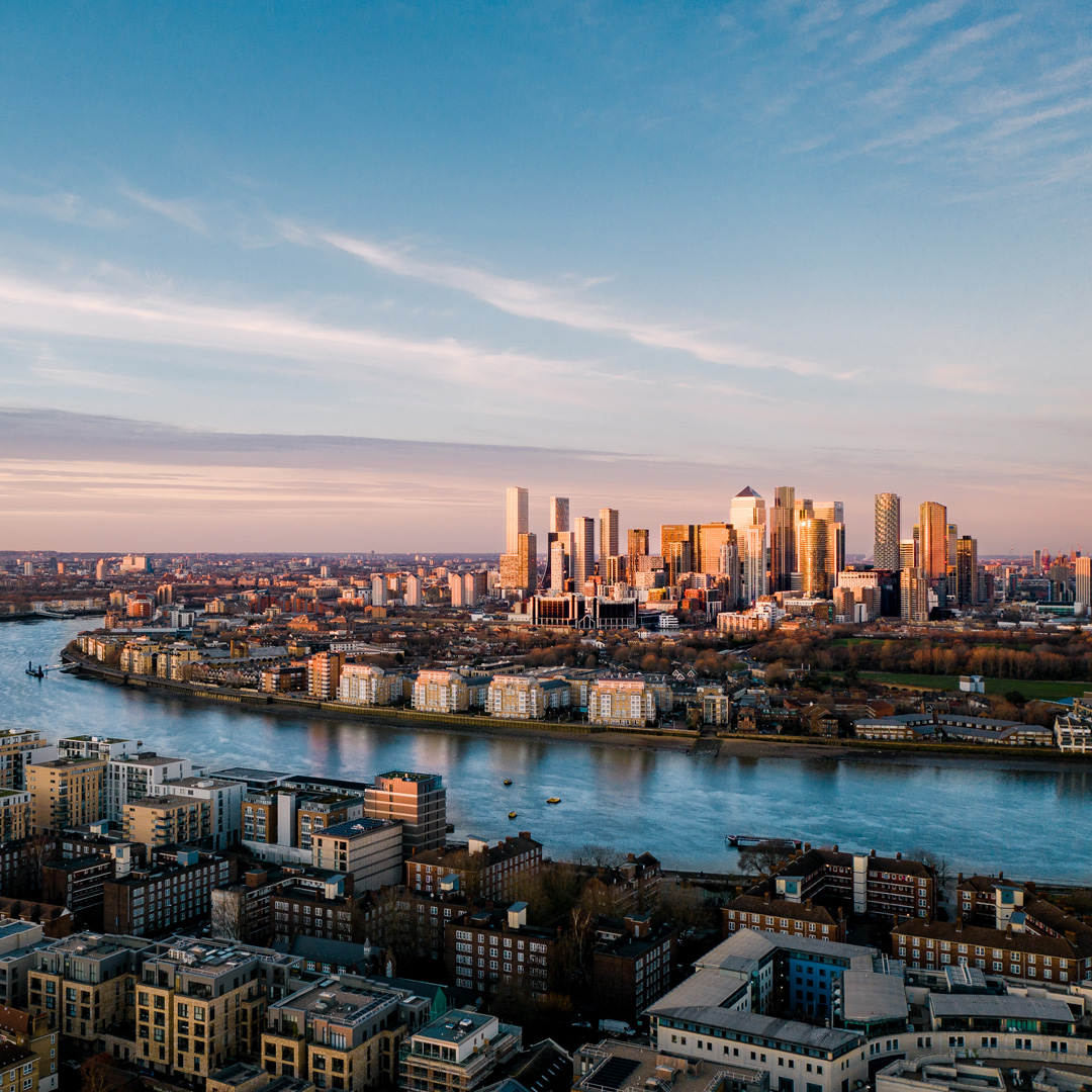 Aerial view of a city with a river running through it during sunset. The skyline features numerous modern skyscrapers reflecting the golden light, with residential and commercial buildings in the foreground.