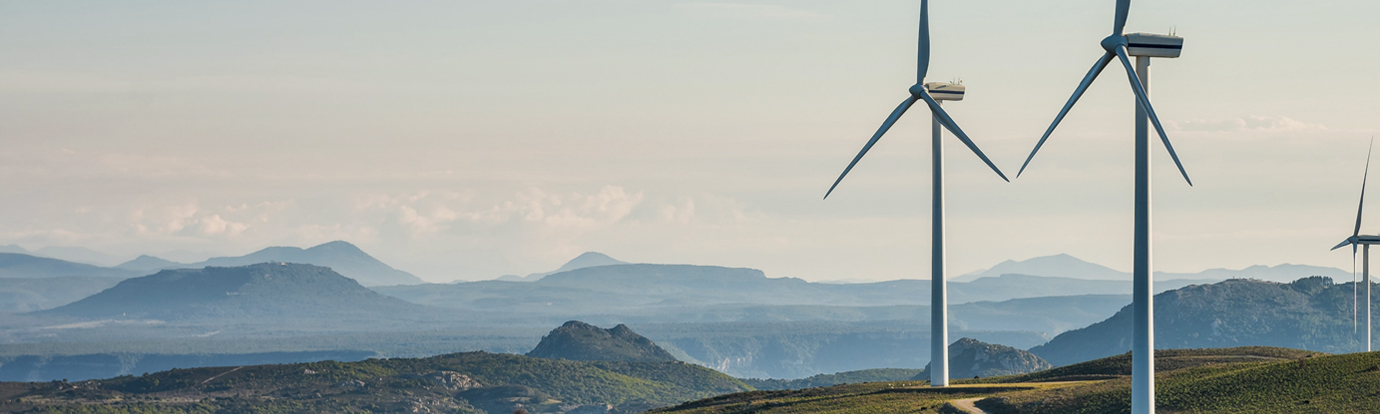Three wind turbines stand on a grassy hill overlooking a vast, hazy mountain range under a pale sky. Renewable energy in a scenic landscape.
