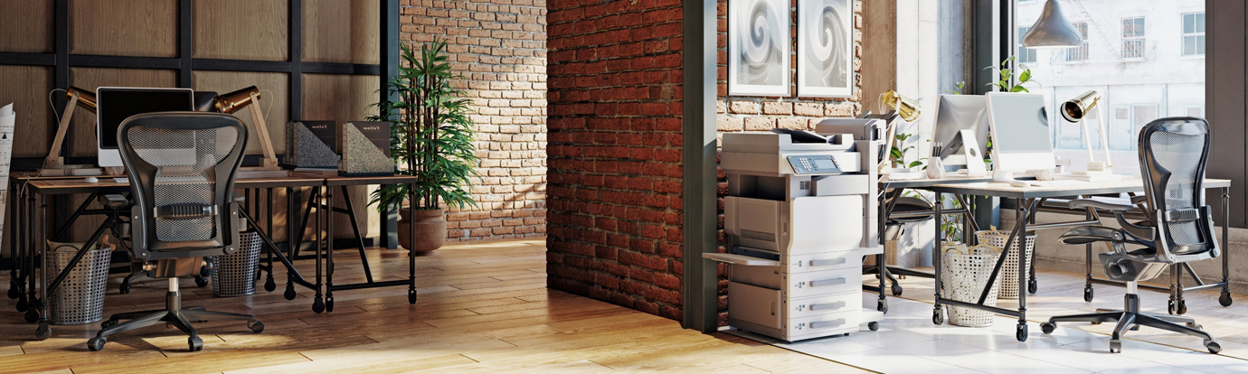 Modern, sunlit office space with two workstations featuring ergonomic chairs, desktop computers, printers, and industrial-style furniture. Exposed brick walls and wood accents create a stylish workspace.