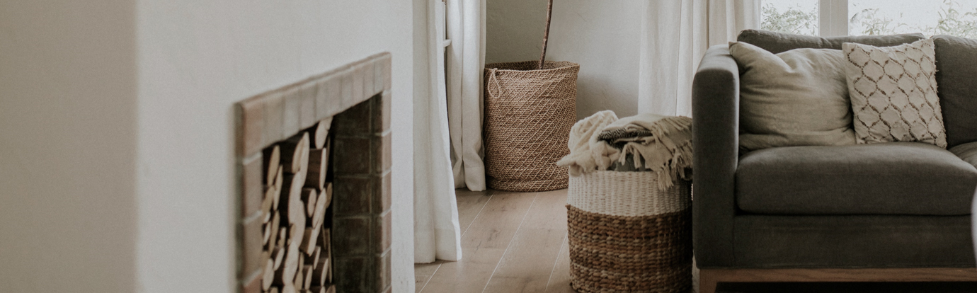A cozy living room scene featuring a gray fabric sofa with beige and off-white textured throw pillows. A woven seagrass basket sits on the floor next to the sofa, containing a folded light beige blanket. Another larger woven seagrass basket stands behind the sofa.
