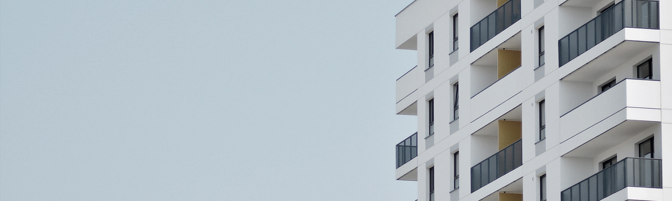 Modern white apartment building exterior with dark grey balconies against a pale blue sky.