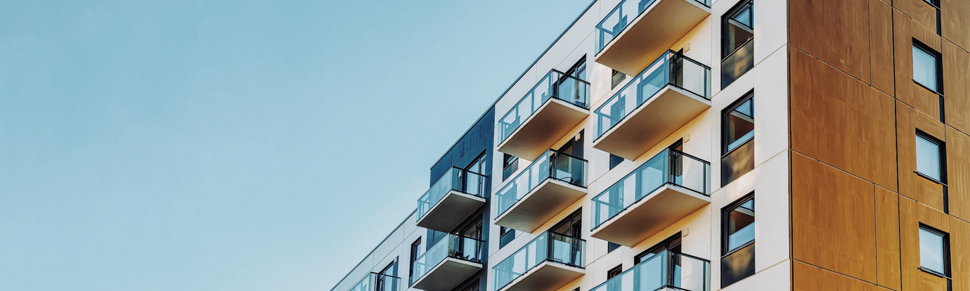 Modern apartment building with balconies and a mix of white and wood-paneled exterior against a clear blue sky.