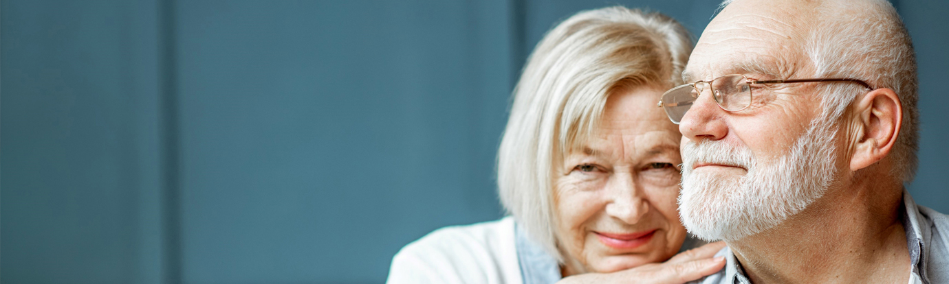 Close-up of a loving elderly couple, the woman with short blonde hair gently embracing the man who is wearing glasses. They are positioned against a muted blue background.