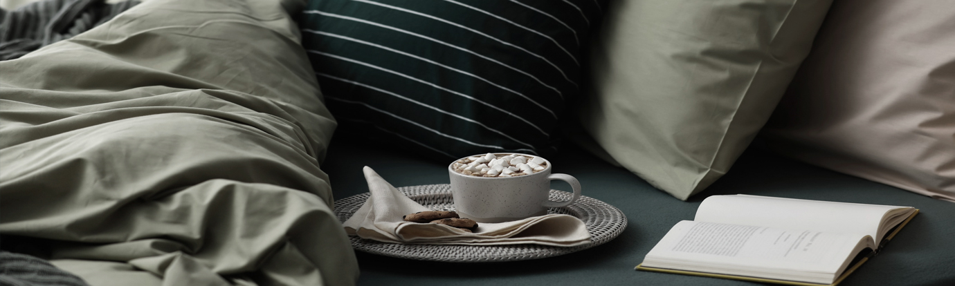 A Cozy hotel room bed: a mug of hot chocolate with marshmallows, cookies, and an open book rest on a bed with green bedding and pillows.