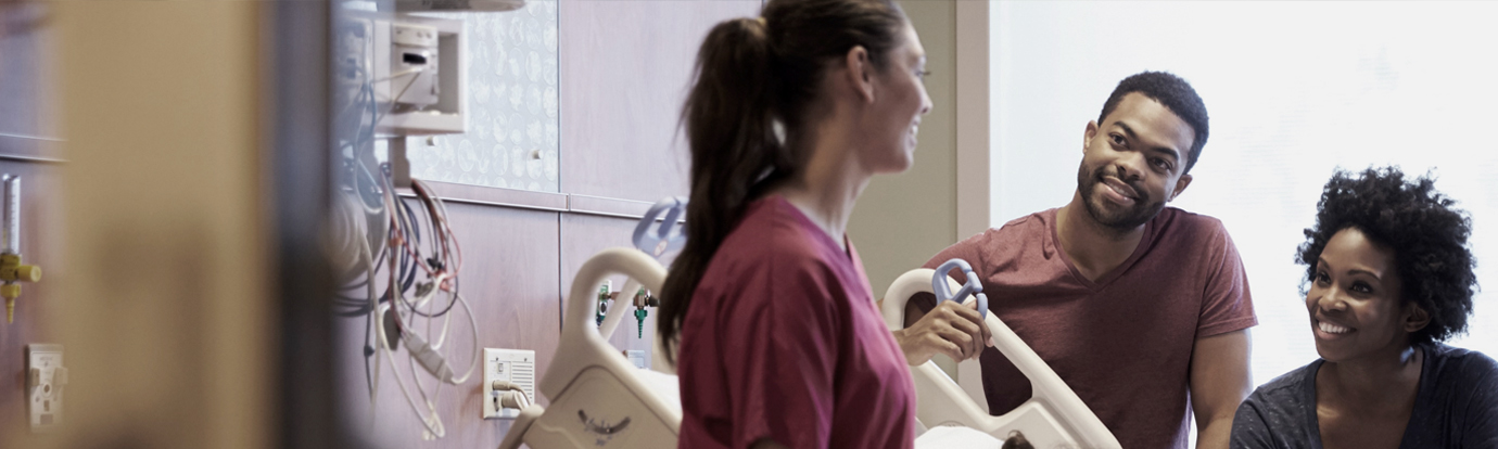 A smiling nurse in a burgundy scrub top speaks with a happy couple in a hospital room. Medical equipment is visible in the background.