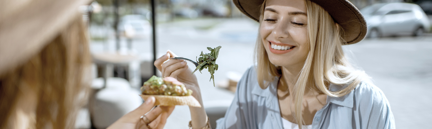 Two women enjoy a meal outdoors. One woman, wearing a brown hat, smiles as she eats a small open-faced sandwich with a side of greens.