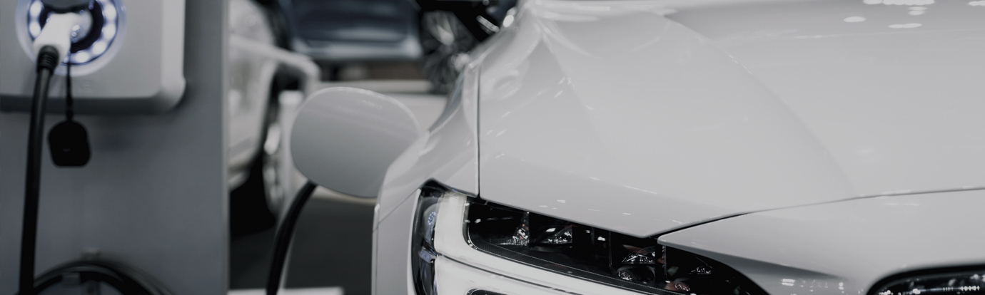 Close-up of a white electric vehicle charging at a charging station. The focus is on the car's front end and the charging cable.