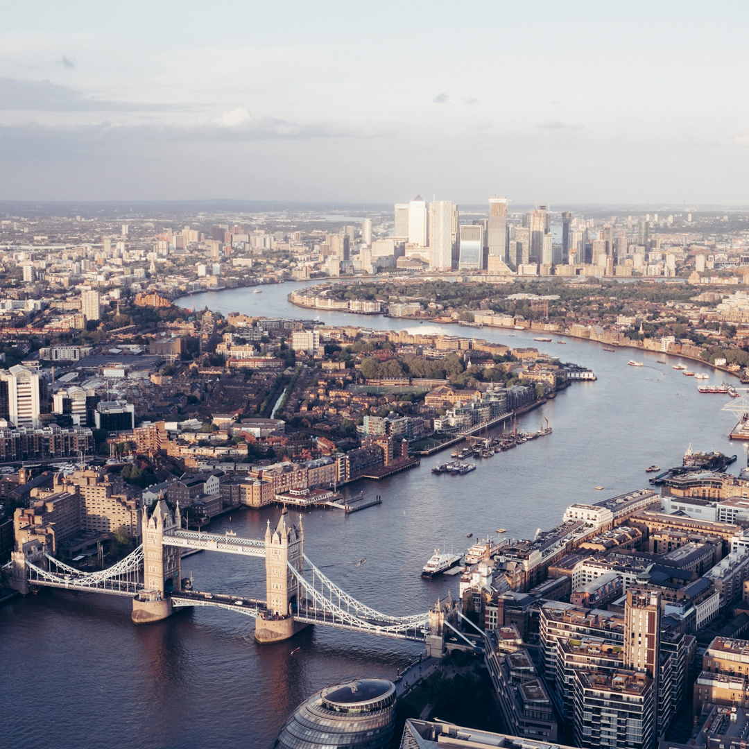 Aerial view of London, England, showcasing the River Thames curving through the city. Tower Bridge is prominently featured in the foreground, its white towers and suspension bridge spanning the river.