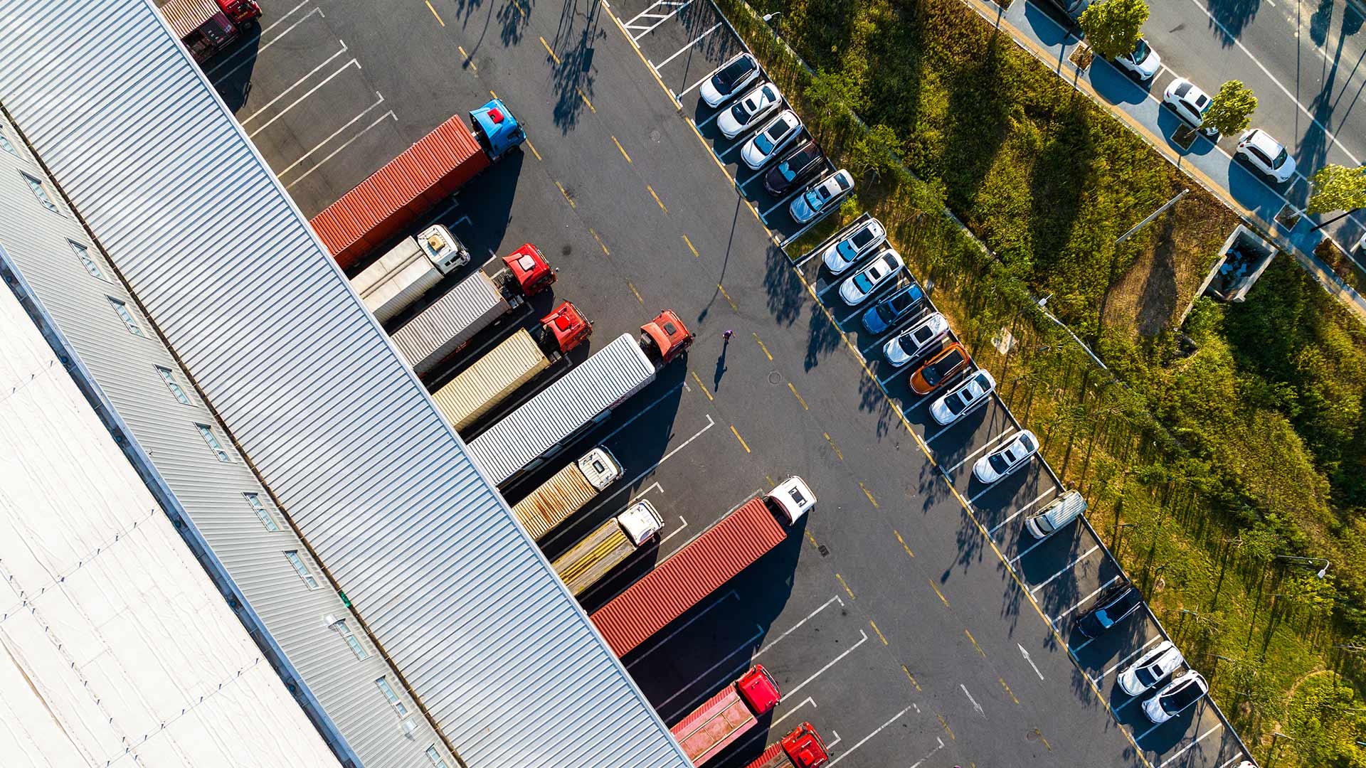 Aerial view of a logistics center with trucks and containers parked in a parking lot. On the right side are several passenger cars, surrounded by greenery and trees. A person walks through the parking lot.