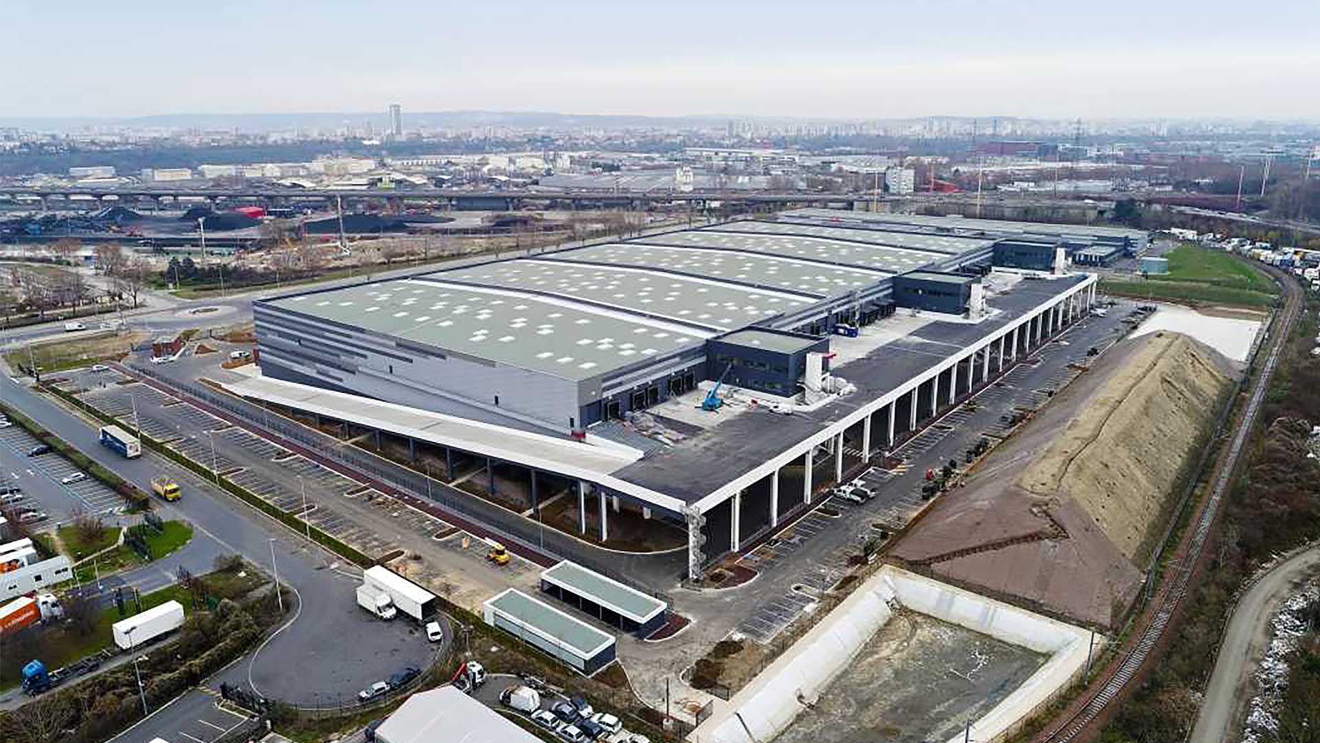 Aerial view of a large distribution center with multiple loading docks, parking lots, and urban environment in the background.