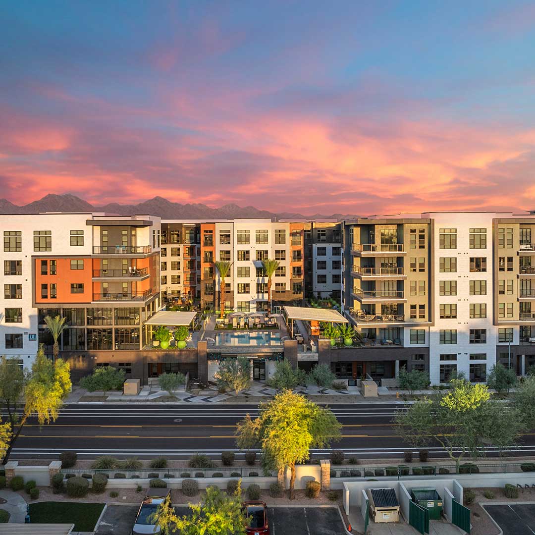 Modern apartment complex at sunset with colorful facades, large windows, balconies, a courtyard pool, parked cars, and mountains in the background.