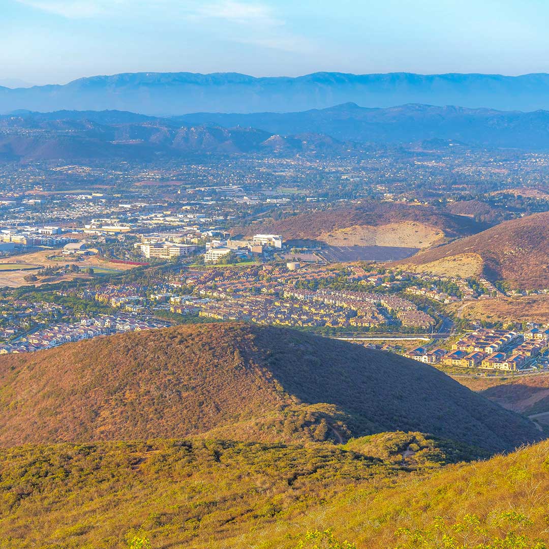 A panoramic view of a suburban area nestled among rolling hills, with a backdrop of distant mountains under a clear sky